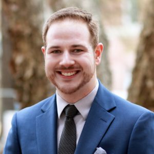 Montgomery_David A man with short light brown hair and a beard is smiling, wearing a blue suit, light shirt, and dark tie—standing outdoors in front of blurred trees, he exudes confidence often seen in accomplished graduate students.