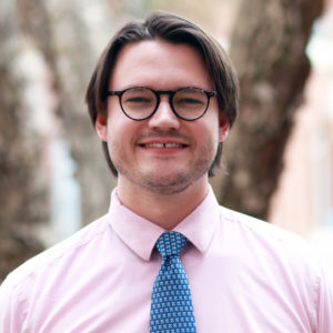 Stott_Garrick A man with medium-length brown hair, glasses, and a short beard smiles while wearing a pink shirt and patterned blue tie, standing outdoors among trees—a look often seen among graduate students.