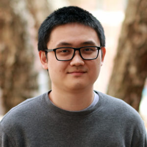 Sun_Kevin A young graduate student with short black hair and glasses, wearing a gray sweater, stands outdoors in front of blurred trees.