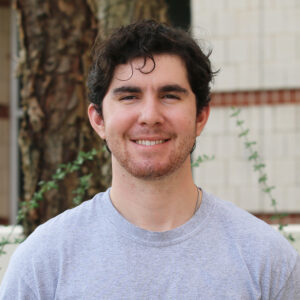 Chris_Esselman A young man with dark curly hair and a short beard, wearing a light gray t-shirt, stands outdoors in front of a tree and a light-colored brick wall, smiling at the camera—typical of many graduate students enjoying campus life.