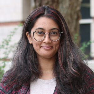 Nikitha_Sundaresha A graduate student with long dark hair and glasses, wearing a sweater, stands outdoors in front of greenery and a blurred building.