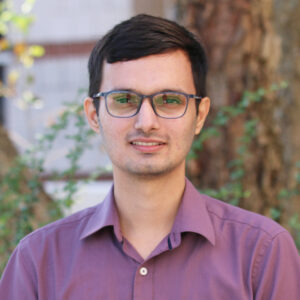 Prawesh_Bhattarai A man with short dark hair and glasses, wearing a purple button-up shirt, stands outdoors in front of a tree and greenery, resembling many graduate students enjoying a break from their studies.