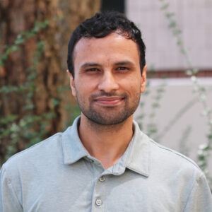 Rajan_Kandel A man with short dark hair and a trimmed beard, wearing a light gray collared shirt, stands outdoors in front of a tree and plants, embodying the focused demeanor often seen in graduate students.