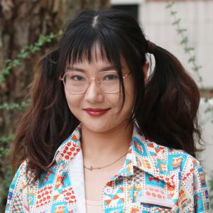 Ying_Xiang A graduate student with glasses and dark pigtails is smiling, wearing a colorful patterned shirt, standing outdoors near a tree.