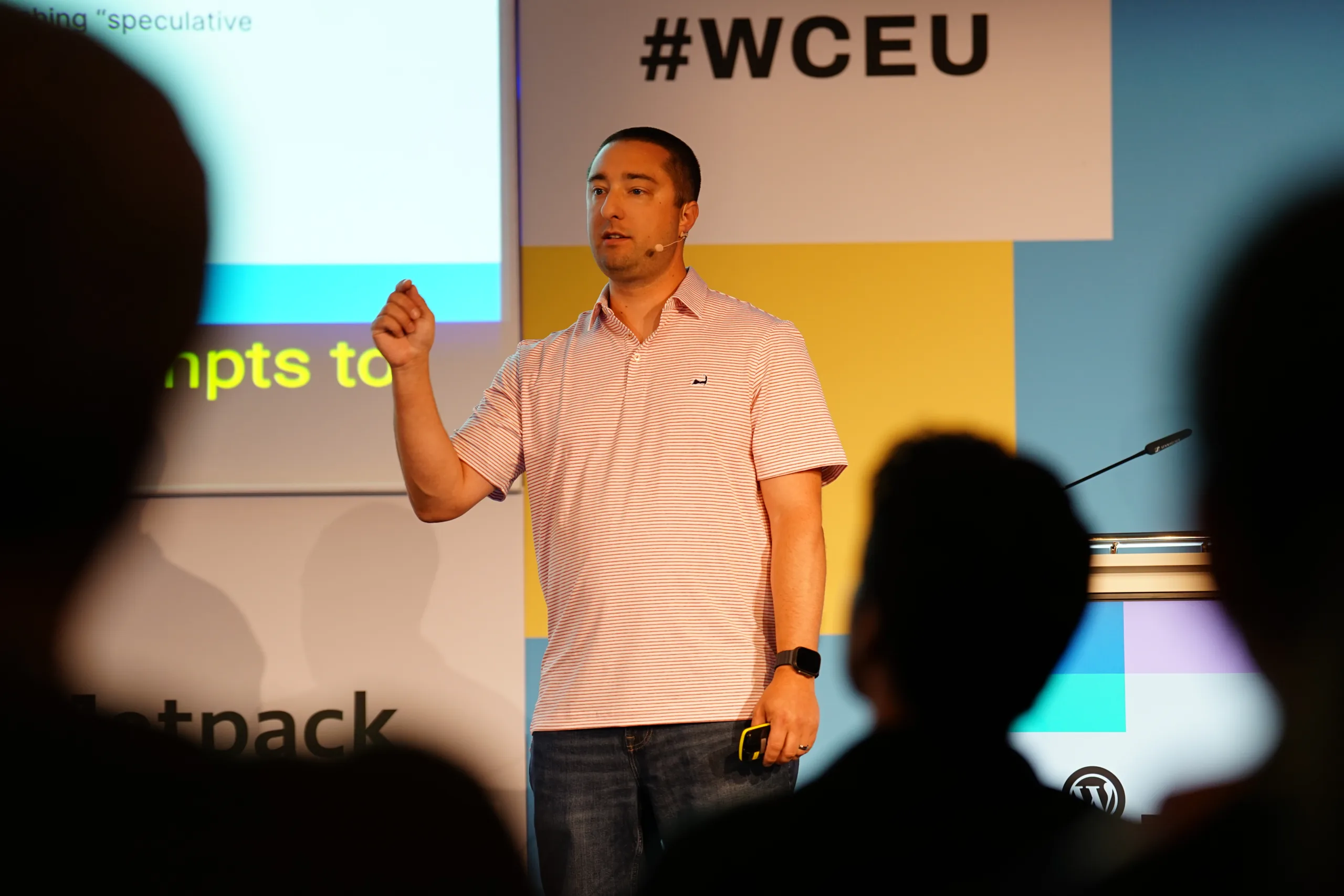 Jonathan Desrosiers speaking on stage at WordCamp Europe 2025. Several silhouettes of attendees are visible in the foreground.