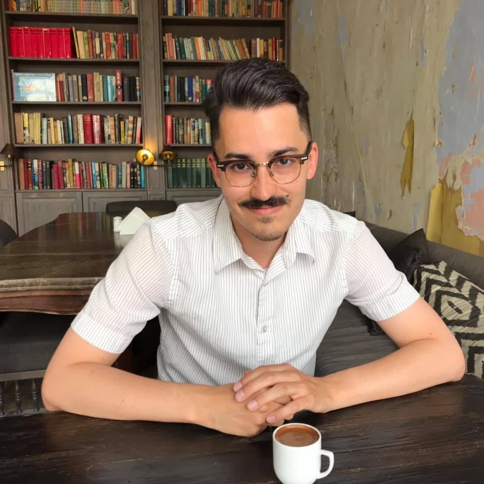 Man with mustache sits with hands clasped in front of a cup. He has glasses.