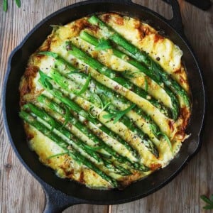 A cast-iron skillet filled with asparagus frittata on a wooden table with a fork on the side.