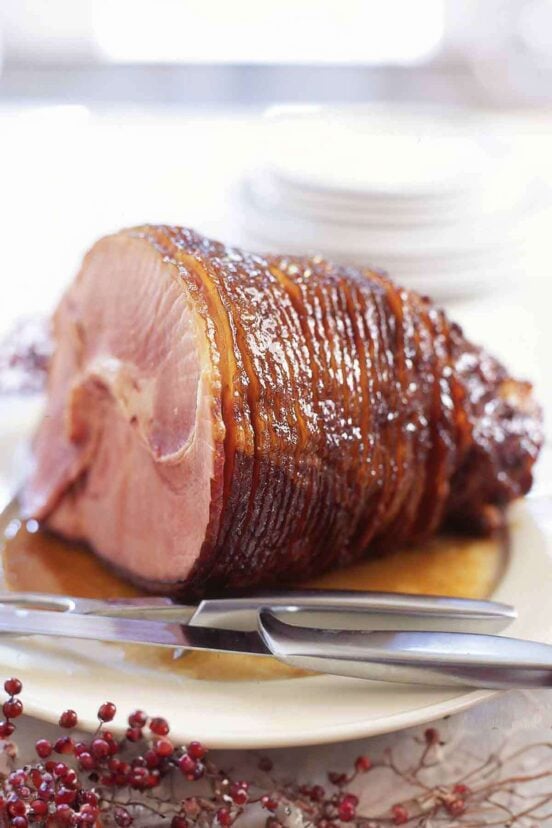 A glazed spiral cut ham on a white platter with a carving knife and fork nearby.