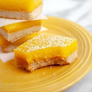 Yellow plate with four lemon bars sprinkled with powdered sugar, one with a bite take out of it.