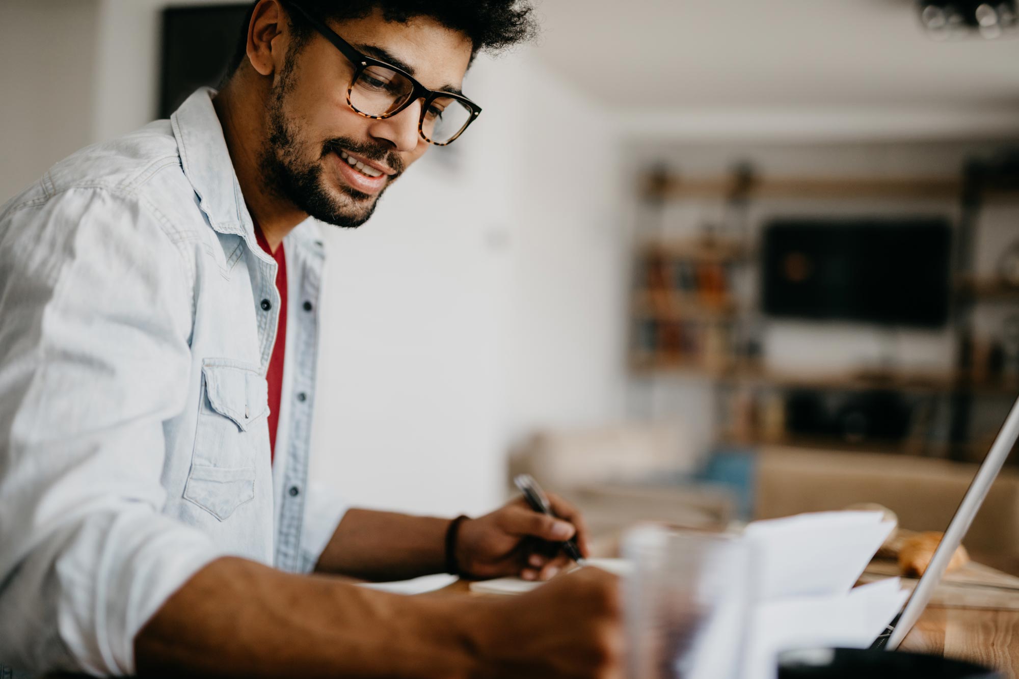 A person works from a table at home, taking notes and working in front of a laptop on a table.