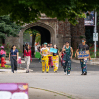 Faculty Representatives walking in front of the arch holding signs that say ' FREE COMPLIMENTS' and 'LOST?'.