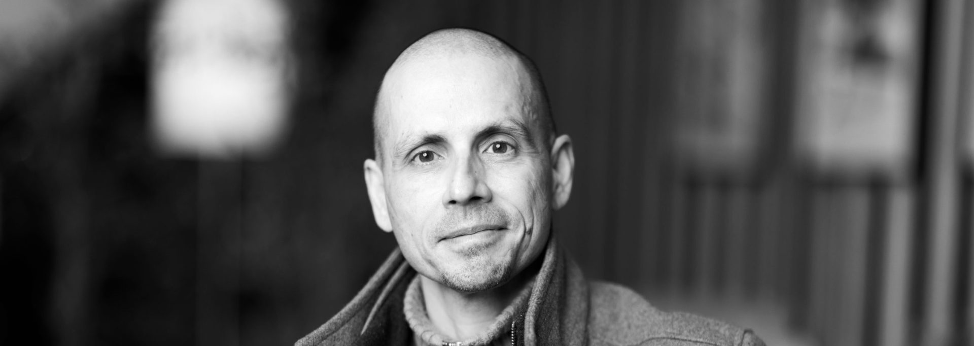Black and white photo of Marc. He is bald and has light colored skin. He is wearing a sweater and wool coat. He seems to be sitting in a room that is blurred in the background.