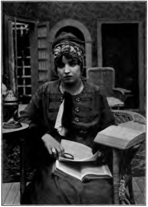 MadelynMack_books.png Woman seated in a chair, with a book on a table at her left side, and another on her lap. She is holding a magnifying glass in her right hand, and looking into the camera.