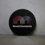 Circular black sign with colourful Music Connects logo leaning against a concrete wall