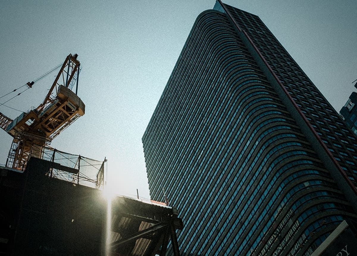 Building and Construction Site Photo of a construction crane beside a tall, curved glass building with sunlight filtering through.