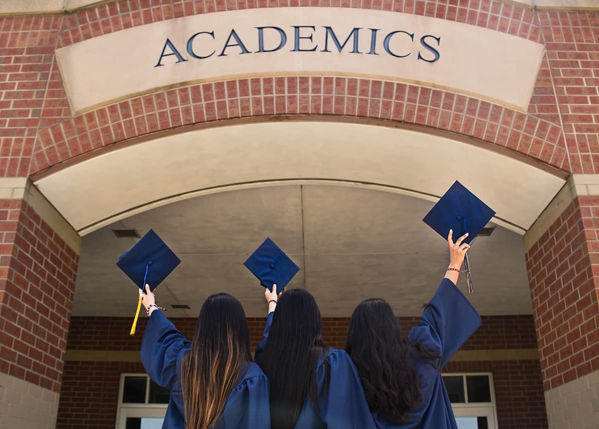 Graduates Celebrating Three graduates in caps and gowns raising their caps in front of a building labeled 'ACADEMICS'.