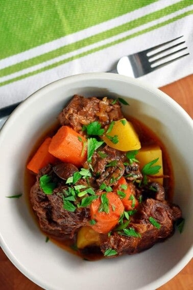 An overhead shot of a white bowl filled with paleo Instant Pot Beef stew with carrots and potatoes. There is a green and white napkin with a fork on top next to the bowl of stew.