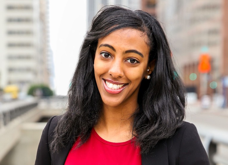Portrait of Black, light-skinned woman with long straight hair. She's wearing a red shirt and black blazer. She's smiling.