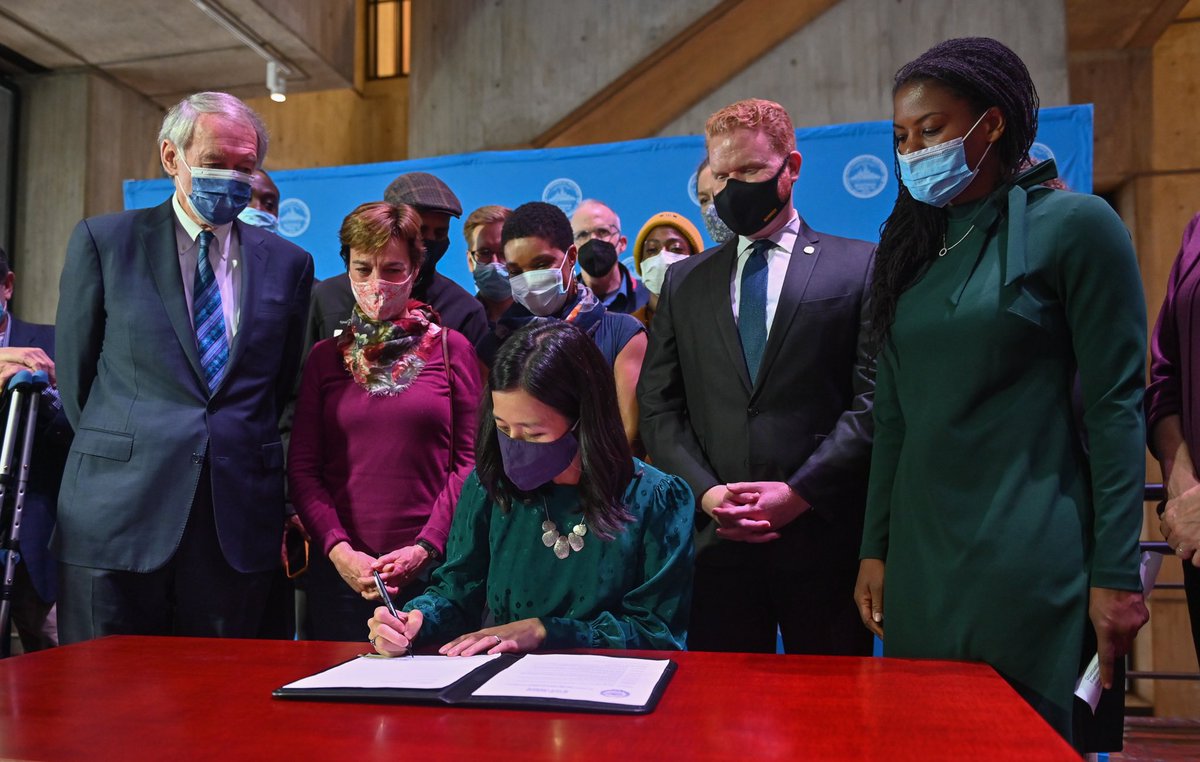Michelle sits at a desk and signs a document as Sen. Ed Markey, Councilor Lydia Edwards, Councilor Matt O’Malley, and environments advocates look on.
