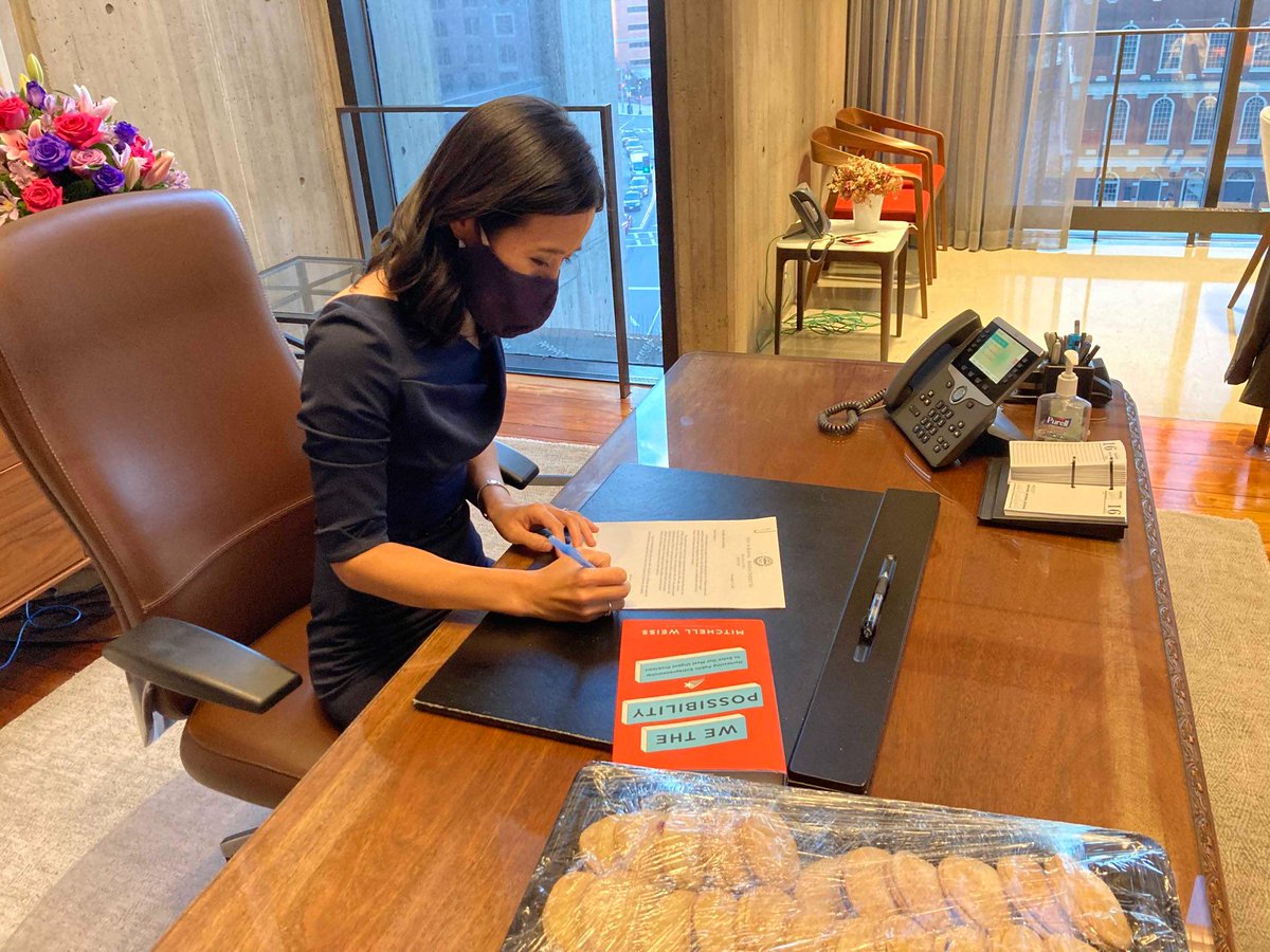 Michelle sits at the Mayor's desk and signs white document.