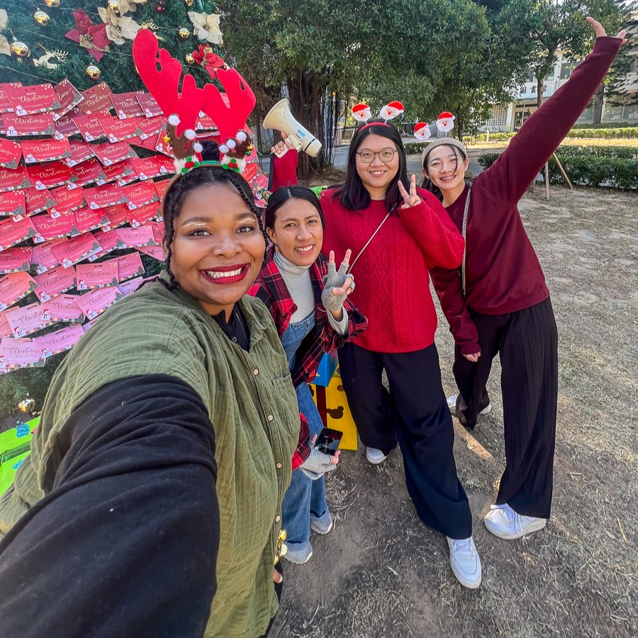 Group of people wearing festive accessories smiling for a selfie in front of a "WISHES" board decorated with holiday themes.