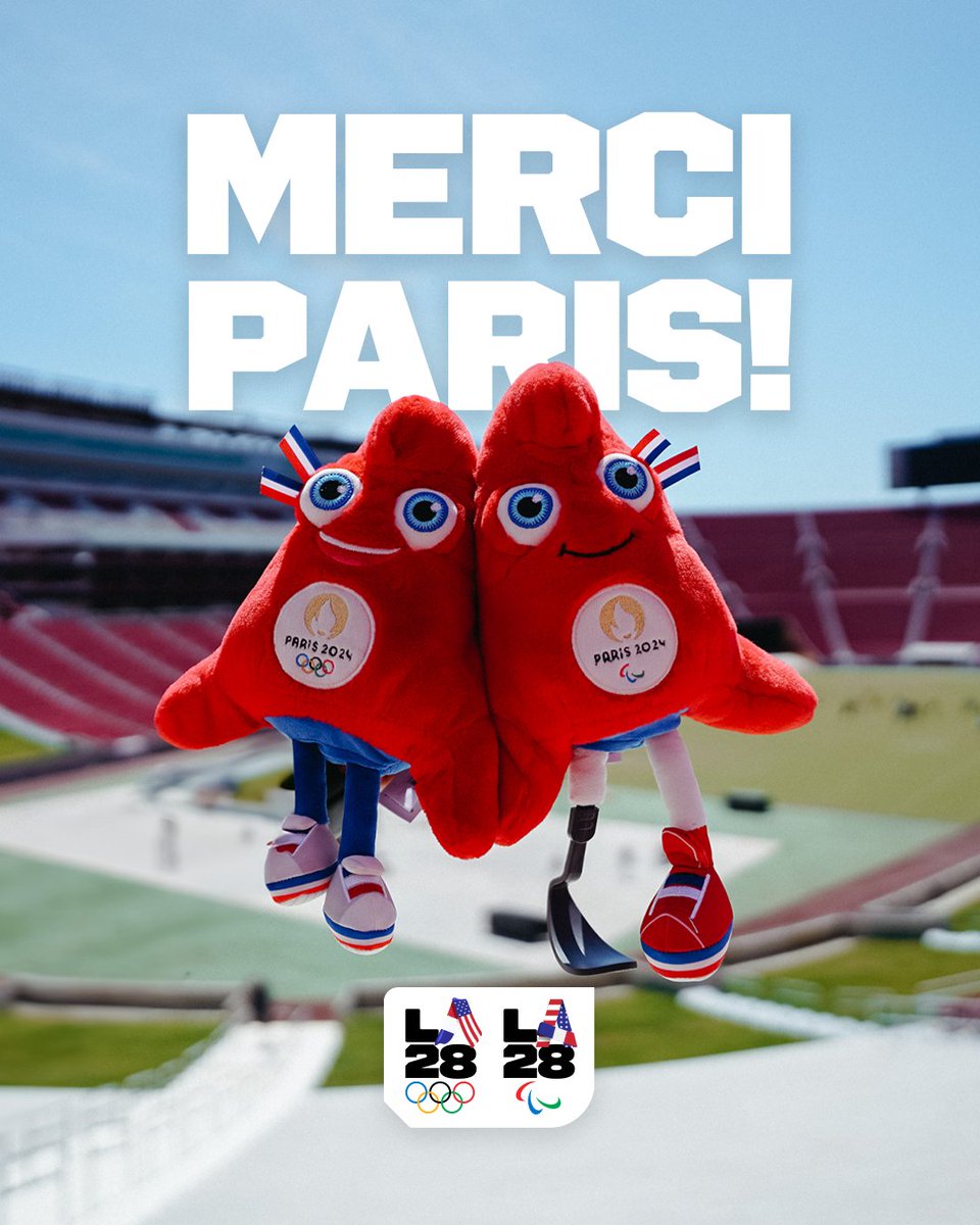 The image shows two Paris 2024 mascots, red plush toys with wide eyes and smiling faces, standing together in front of a sports stadium. They each have the Paris 2024 emblem on their chests, with one mascot having a prosthetic leg. Above them is the bold text “Merci Paris!” in white, expressing gratitude to Paris. Below them, the LA28 Olympic and Paralympic logos are displayed, with American flag elements in the "A" letters. The background features a blurry stadium, likely referencing the transition from Paris 2024 to LA