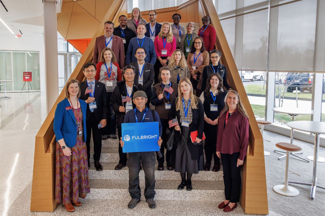 Group of Fulbright program participants standing on stairs in a modern building, smiling at the camera. One person in the front holds a sign with the Fulbright logo.