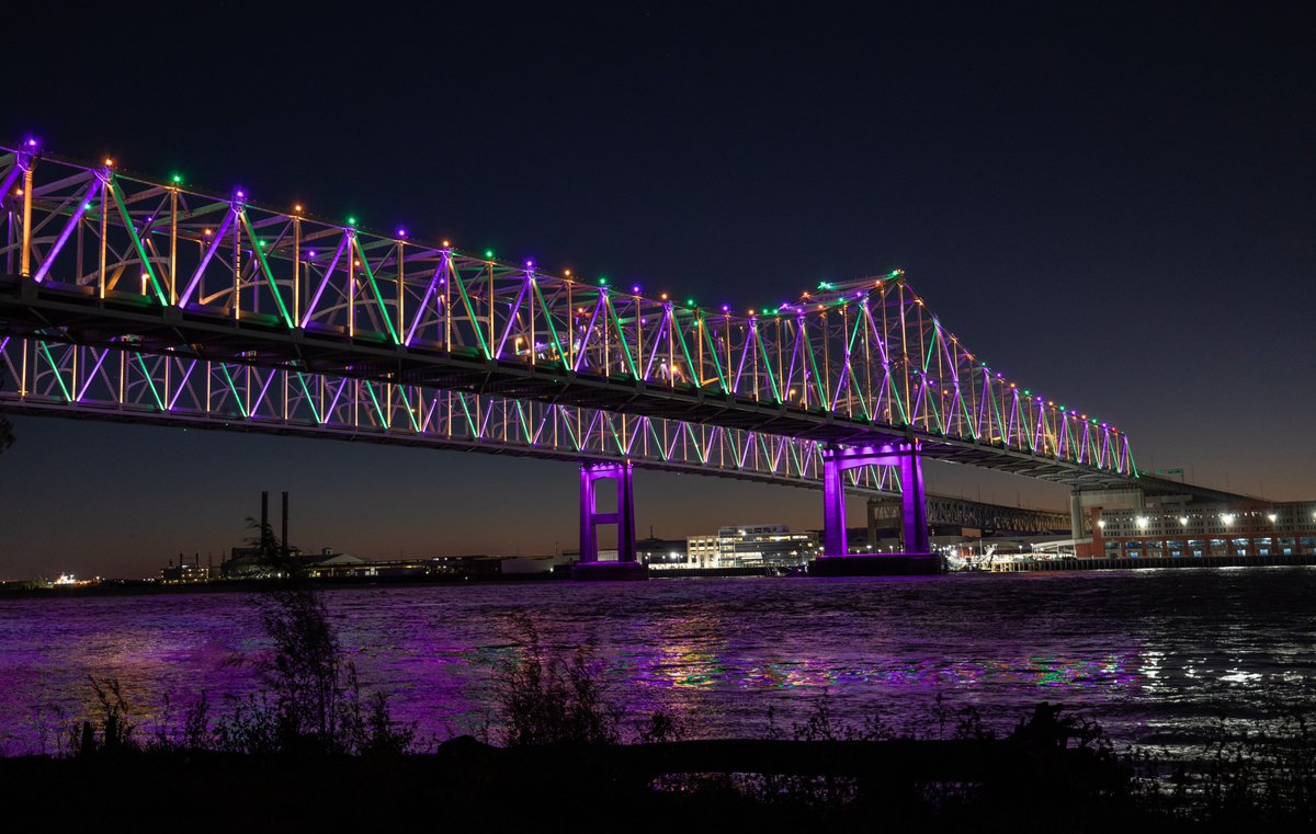 A night view of the illuminated Crescent City Connection bridge over the Mississippi River in New Orleans. The lights are purple, green, and yellow.