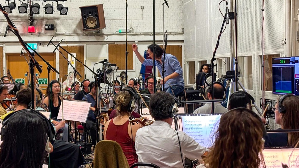A group of musicians in a recording studio are seated with various instruments and sheet music. They are wearing headphones, and microphones surround them. A conductor is standing, gesturing with a baton, leading the orchestra. The room has speakers and sound equipment visible.
