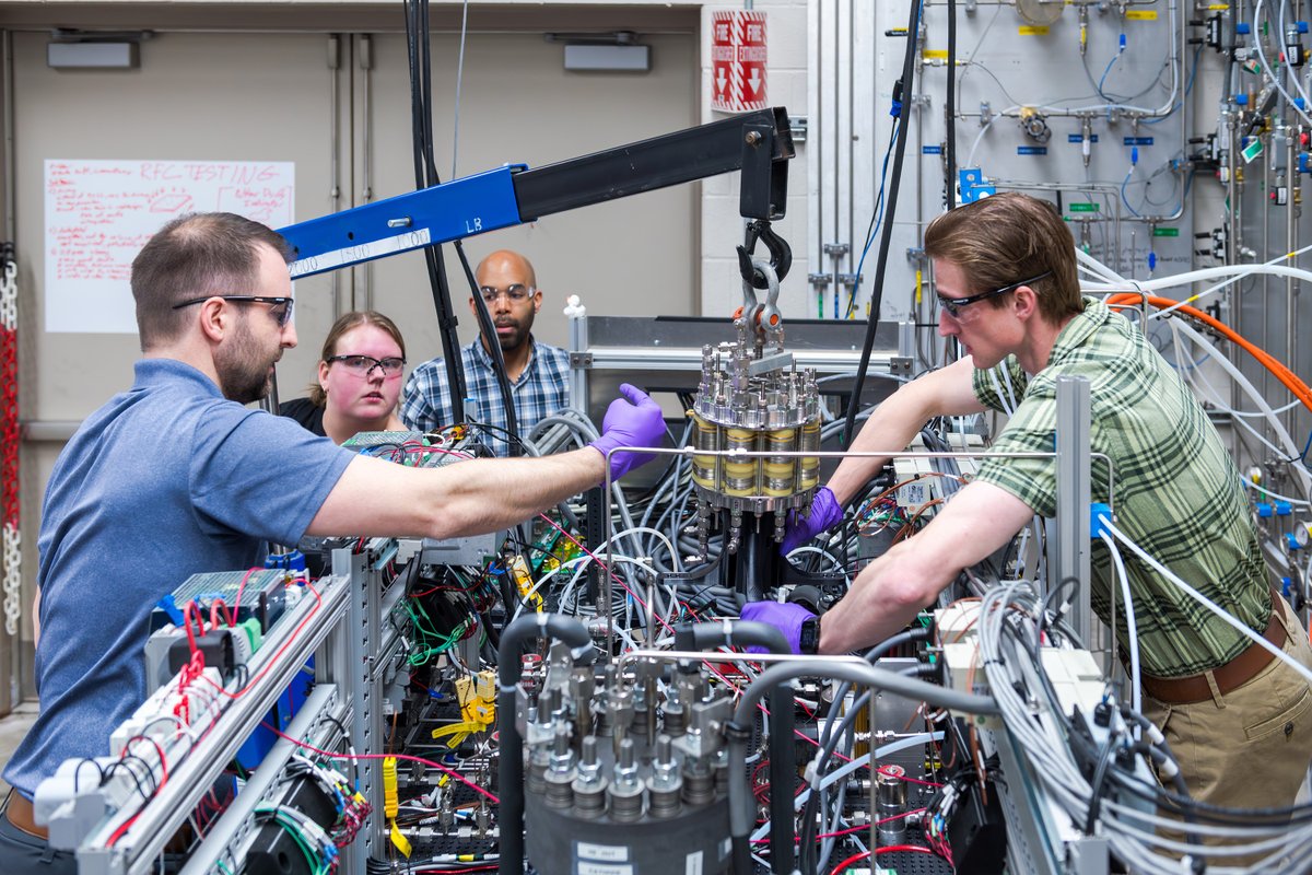 Two researchers in the foreground reach inside a system filled with tubes, wires, and hardware. One researcher points left to direct another researcher on where to place a small fuel cell hanging from a blue crane. Two other researchers look on in the background.\u202F Credit: NASA/Jef Janis