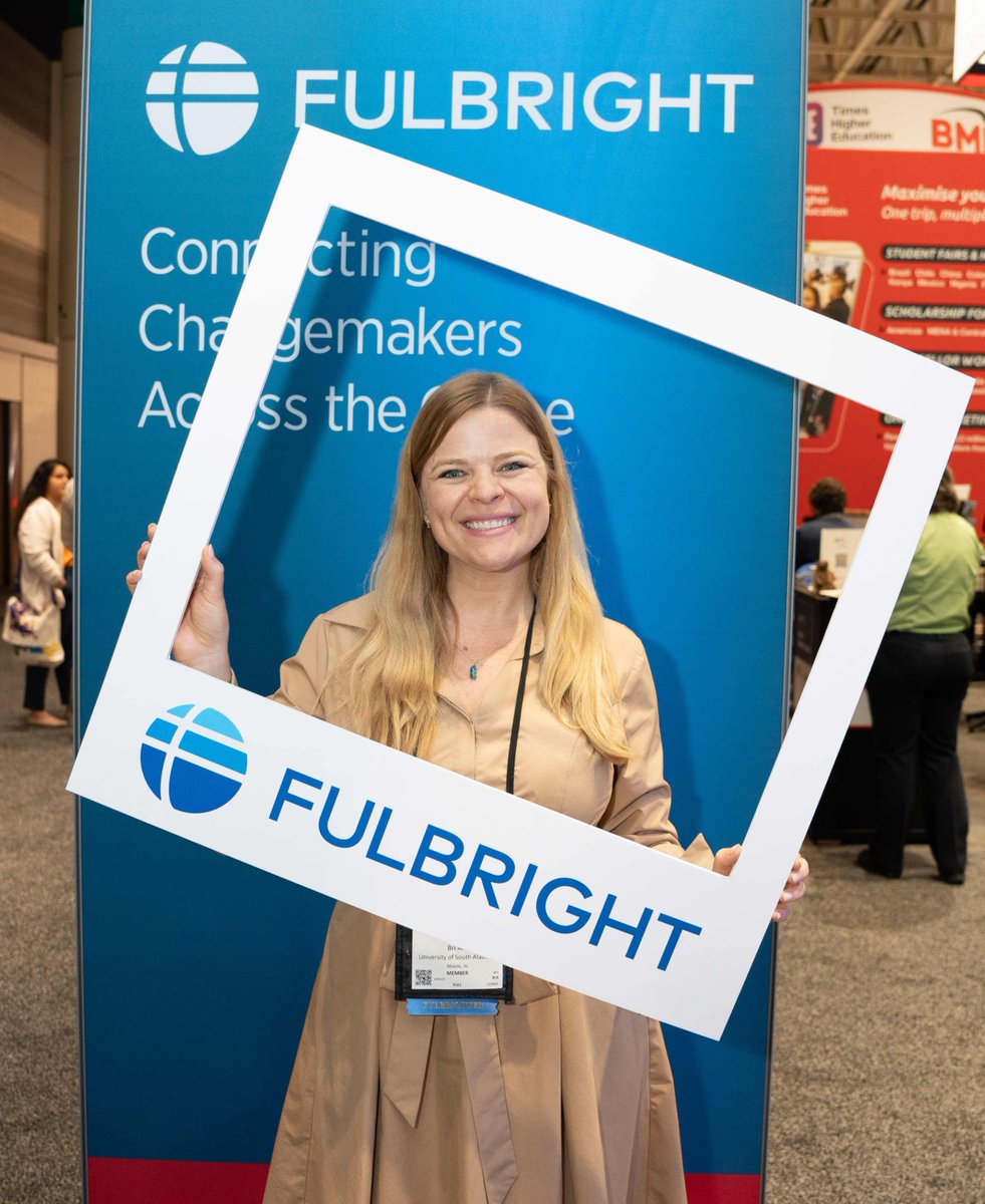 A person smiling while holding a Fulbright-branded frame in front of a Fulbright booth display. The background includes informational signage and blurred figures walking by.