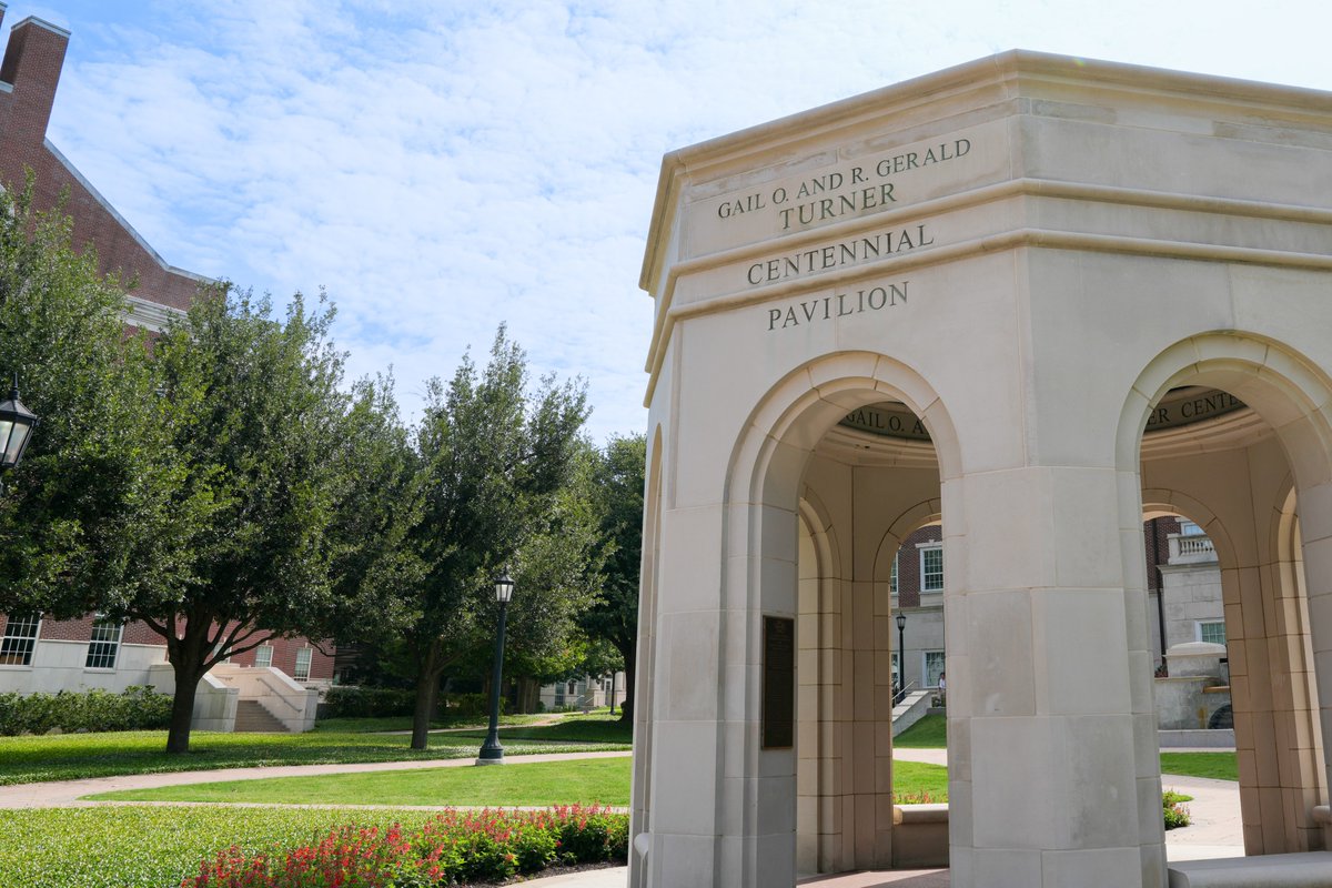 The image shows the Gail O. and R. Gerald Turner Centennial Pavilion, an architectural structure with arches, located on a grassy area surrounded by trees and flowers. A brick building is visible in the background.