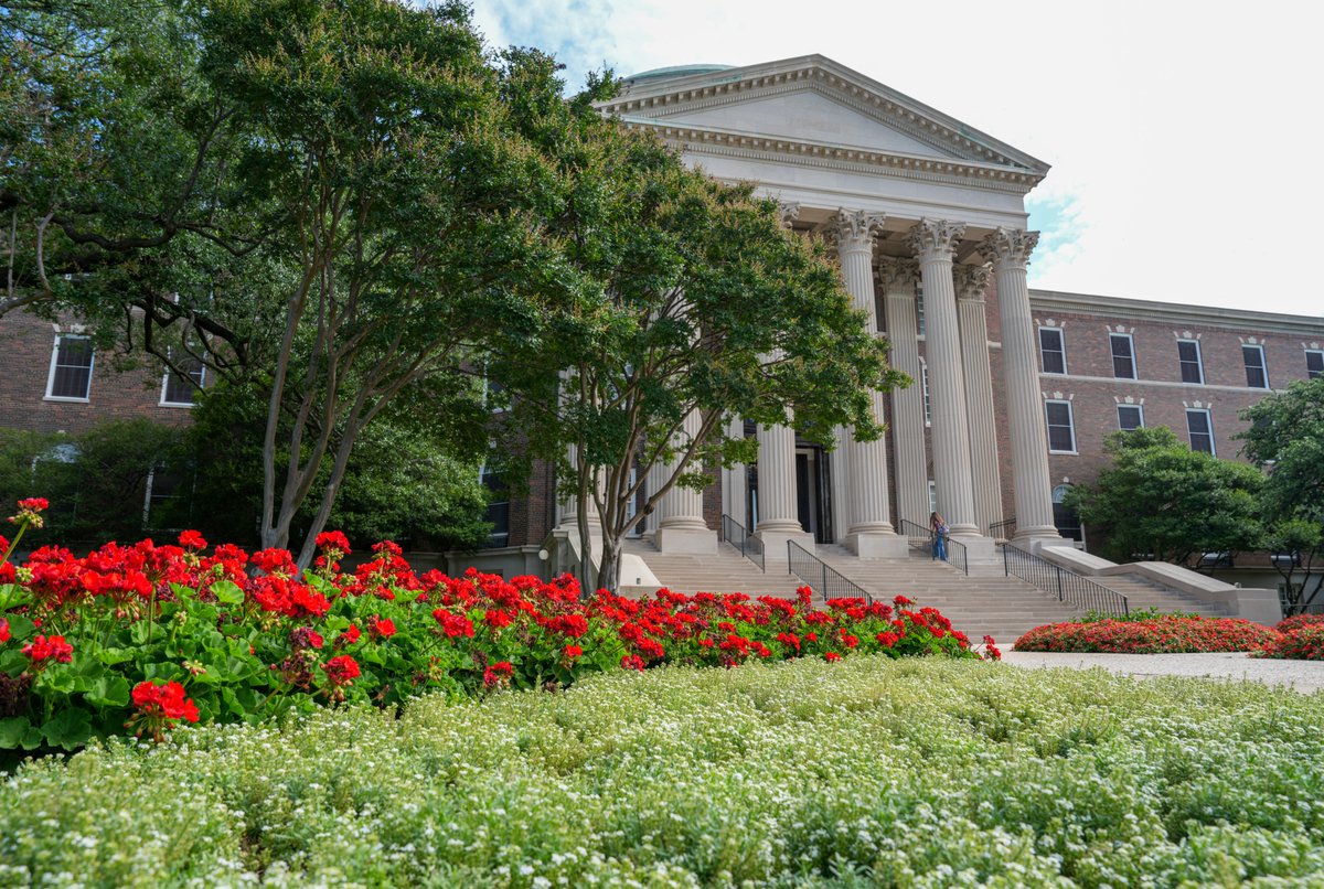 The image shows the exterior of Dallas Hall at SMU, a large classical-style building with tall white columns at the entrance, surrounded by neatly landscaped gardens featuring red and white flowers beneath a partly cloudy sky.