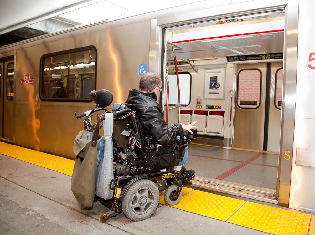 Une personne en fauteuil roulant électrique monte à bord d’une rame de métro accessible depuis le quai d’une station doté d’une zone d’embarquement de plain-pied et d’une bande de sécurité podotactile.