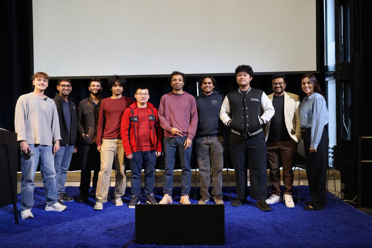 A group of nine individuals is standing on a blue carpet in front of a large screen. They are casually dressed and smiling, with one person wearing a jacket featuring the "Aether" logo.