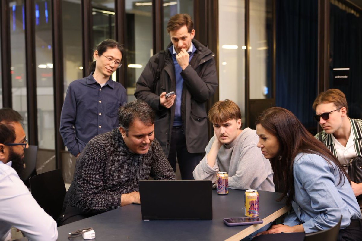 A group of people gathered around a table, looking at a laptop. One person in a dark shirt is seated and focused on the screen. Others stand or sit close by, intently observing, while some hold drinks. The setting appears to be a modern, indoor environment with large windows.