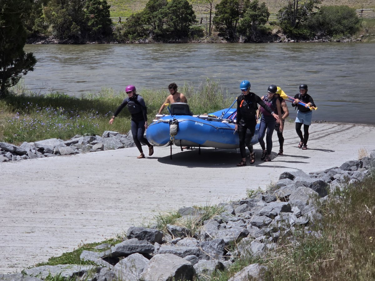 Group of five people carrying a blue inflatable raft toward a river on a rocky path surrounded by greenery.
