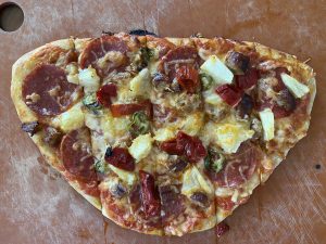 Hand-tossed thin-crust pizza on cutting board, with cheese, pepperoni, sausage, goat horn pepper, pineapple, and jalapeño