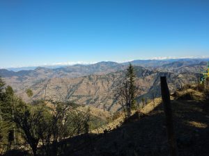A scenic mountain landscape with a clear blue sky. The foreground features some trees and rocky terrain, while distant snow-capped peaks are visible on the horizon.