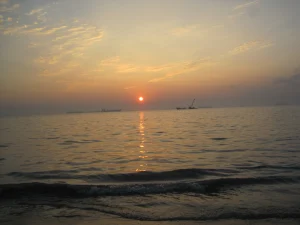 A serene seascape at sunset, showing the sun dipping below the horizon, casting a warm orange glow across the sky and shimmering on the water’s surface. Silhouetted ships are visible in the distance.