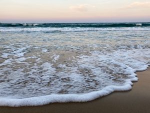 A serene beach scene featuring gentle waves lapping against the sandy shore. The water glistens under soft daylight, and a gradient of pastel colors fills the sky, indicating either sunrise or sunset.