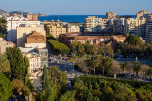 The city of Malaga taken from a hillside. The foreground is made up of green trees and the La Malagueta bullring sits to the right of the frame set amongst modern residential tower blocks and a line of trees. The coastline and sea is visible as it forms the horizon with a deep blue sky.