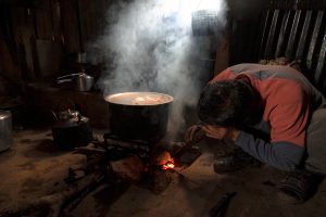 A person crouching next to a wood-powered stove in a dimly lit room, tending to a fire beneath a large pot with steam rising.