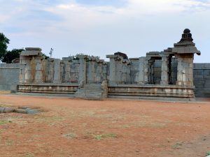 Ancient stone temple ruins with columns, situated on a sandy terrain under a partly cloudy sky. The structure features detailed carvings and stone steps leading up to the platform.