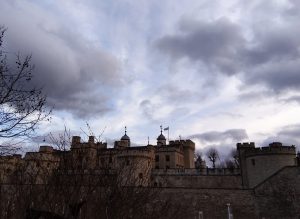A historic stone fortress with towers and battlements silhouetted against a cloudy sky. Leafless trees are visible in the foreground.