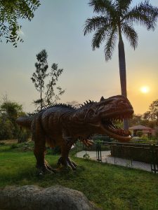 A large dinosaur statue with an open mouth stands in a grassy area surrounded by trees, including a tall palm tree, under a setting sun.