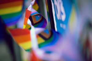 A close-up shot of fluttering rainbow and Progress Pride flag bunting captures vibrant colors in motion. The shallow depth of field creates a dreamy blur, with hints of signage and background color.