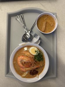 Tray with curry noodle soup topped with shrimp, boiled egg, and cilantro, served with a side of thick reddish-brown sauce.