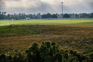 A wide view of a golf course taken during the day, featuring a partially cloudy sky with rays of light streaming through. In the foreground, there is a patch of grass with some dry areas and a cluster of cacti. In the background, the green fairway stretches across the scene, with flags hinting at various holes.