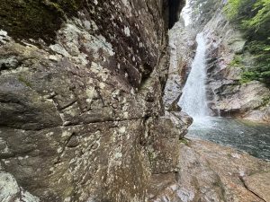 A rock wall at the base of Glen Ellis Falls, New Hampshire, carved with ‘H.A. Seavey and A.P. Thompson, 1876,’ with the waterfall flowing into the river below.